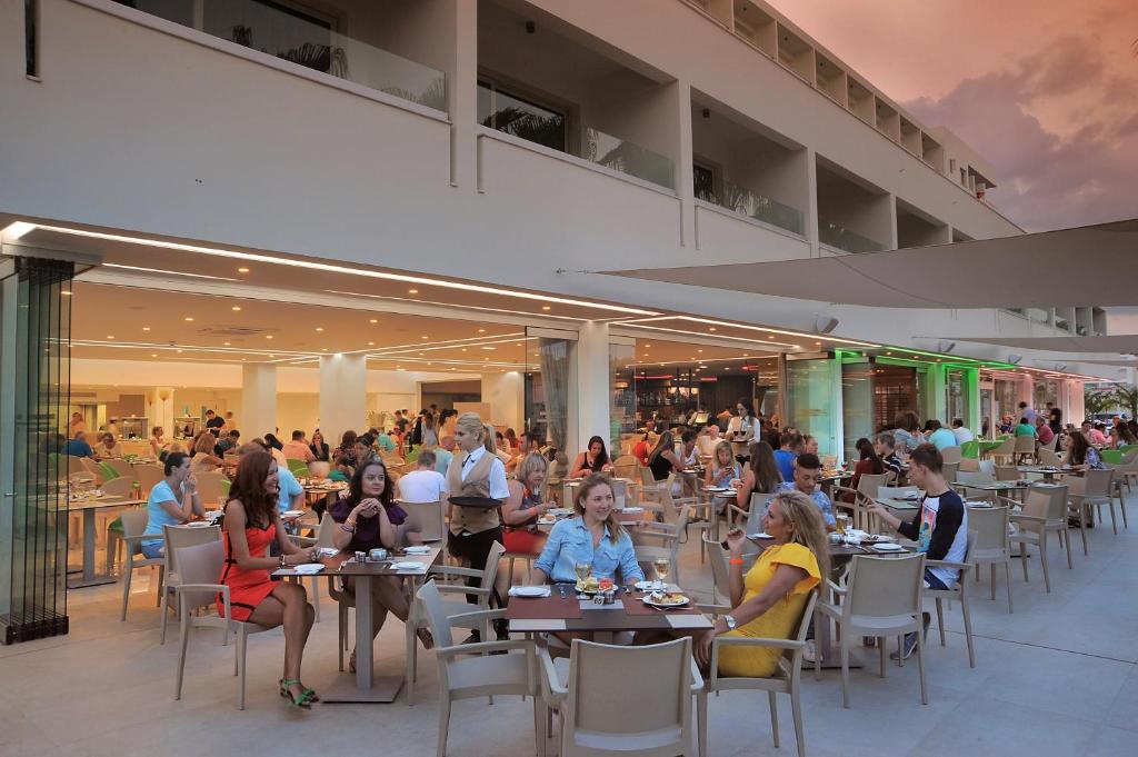 Āra restorāns ar daudzām viesībām, kur cilvēki bauda maltītes, sēžot pie galdiem. Interjers ir gaišs un mūsdienīgs, ar nakts apgaismojumu un relaksētu atmosfēru.