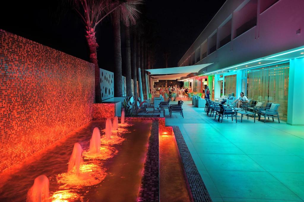 Moderns nakts skats uz viesnīcu ar krāsainiem apgaismojumiem un strūklaku, kas rada relaksējošu atmosfēru. Terase ir pilna ar cilvēkiem, kas bauda vakaru.