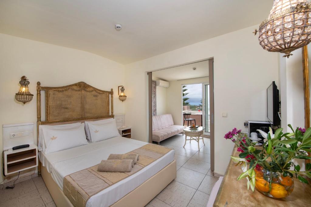 Mūsdienīga viesnīcas istaba ar divguļamo gultu, siltām krāsām un ērtu atpūtas zonu. Loga tuvumā ir gaisīga viesistaba ar piekļuvi balkonam un ziedu kompozīcija uz galda.