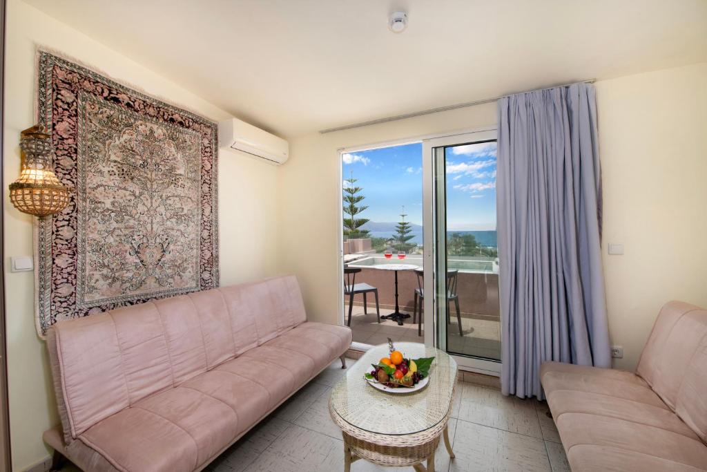 Mūsdienīga viesistaba ar rozā dīvāniem un paklāju pie sienas, kas piedāvā skatu uz jūru caur lieliem logiem. Uz galda novietota augļu kompozīcija, radot mājīgu atmosfēru.