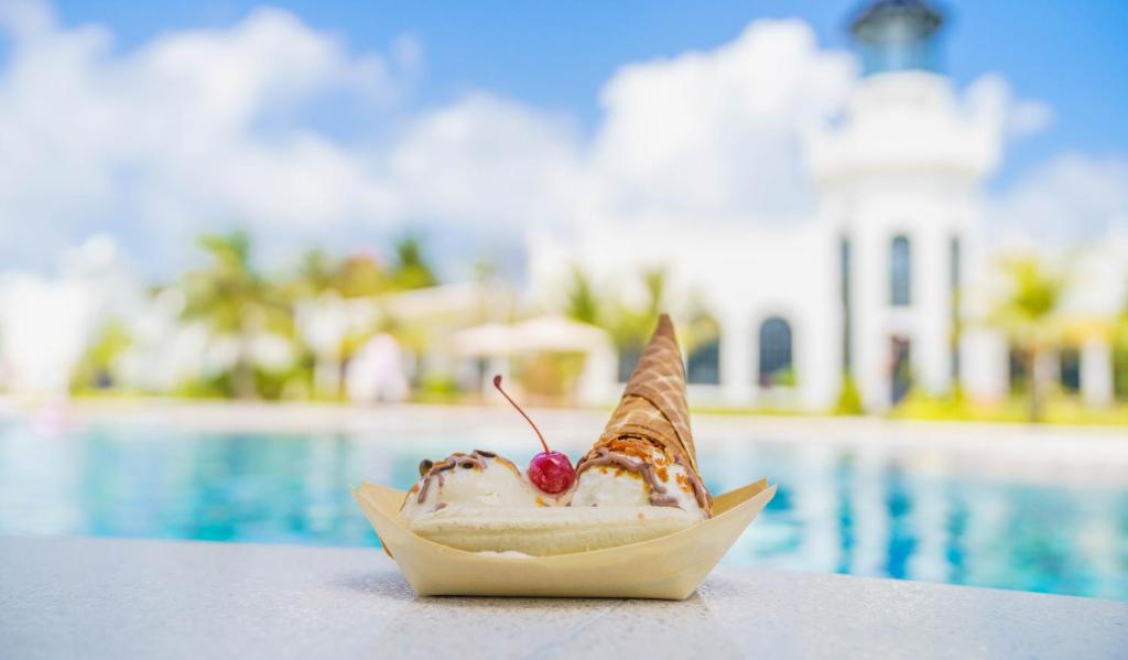 Salda saldējuma vafeļu konuss ar ķirsīti, novietots pie baseina ar palmām un viesnīcas ēku fonā.