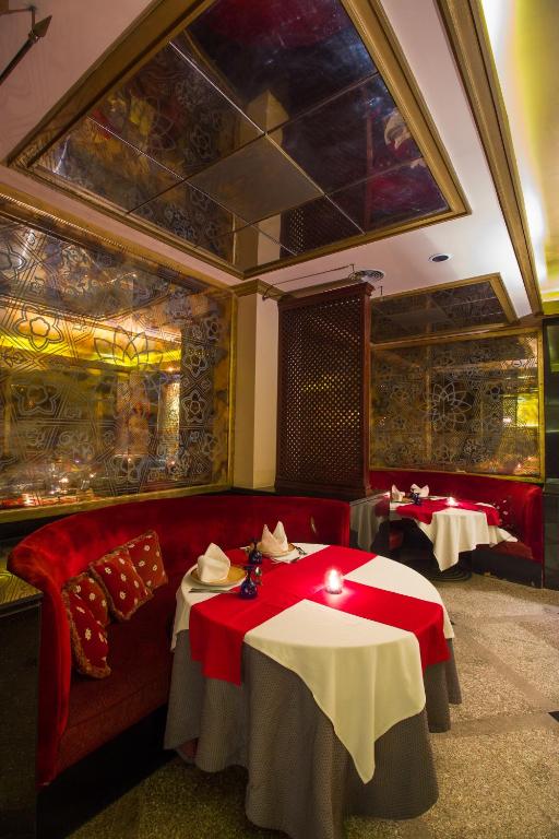 Elegants restorāna interjers ar sarkanu dīvānu un galdiem, klātiem ar baltiem galdautiem, radot intīmu atmosfēru. Spoguļi un dekoratīvie elementi piešķir telpai izsmalcinātību.