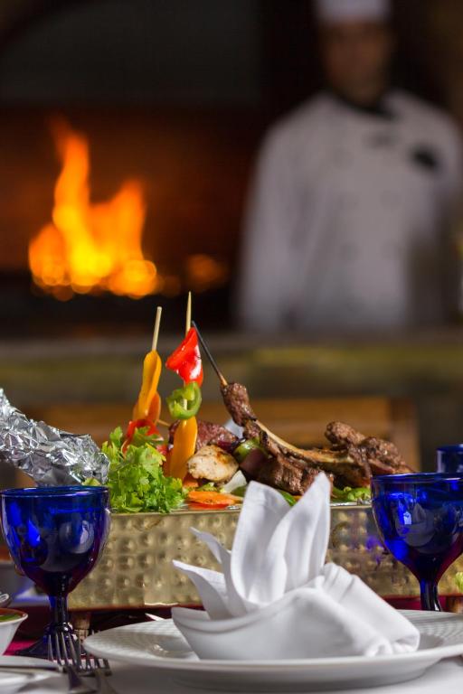 Grilēti gaļas un dārzeņu šašliki uz zelta šķīvja ar zilu trauku fona, kamēr aiz tiem redzams uguns liesmas un šefpavārs.