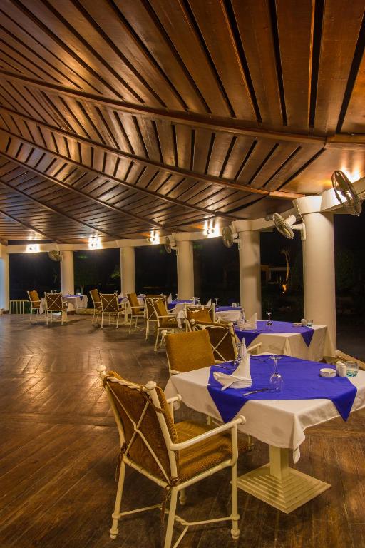 Elegants restorāns ar koka griestiem un apgaismojumu, kurā ir klāti galdi ar ziliem galdautiem un krēsliem. Ideāla vieta romantiskām vakariņām vai draugu sanākšanām.