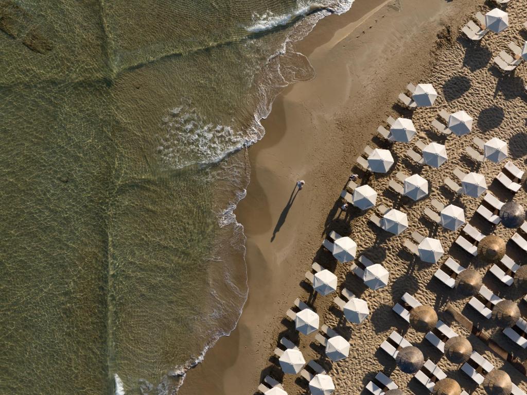 Pludmales skats no augšas ar baltām saulessargiem un cilvēku, kas pastaigājas gar ūdens malu.