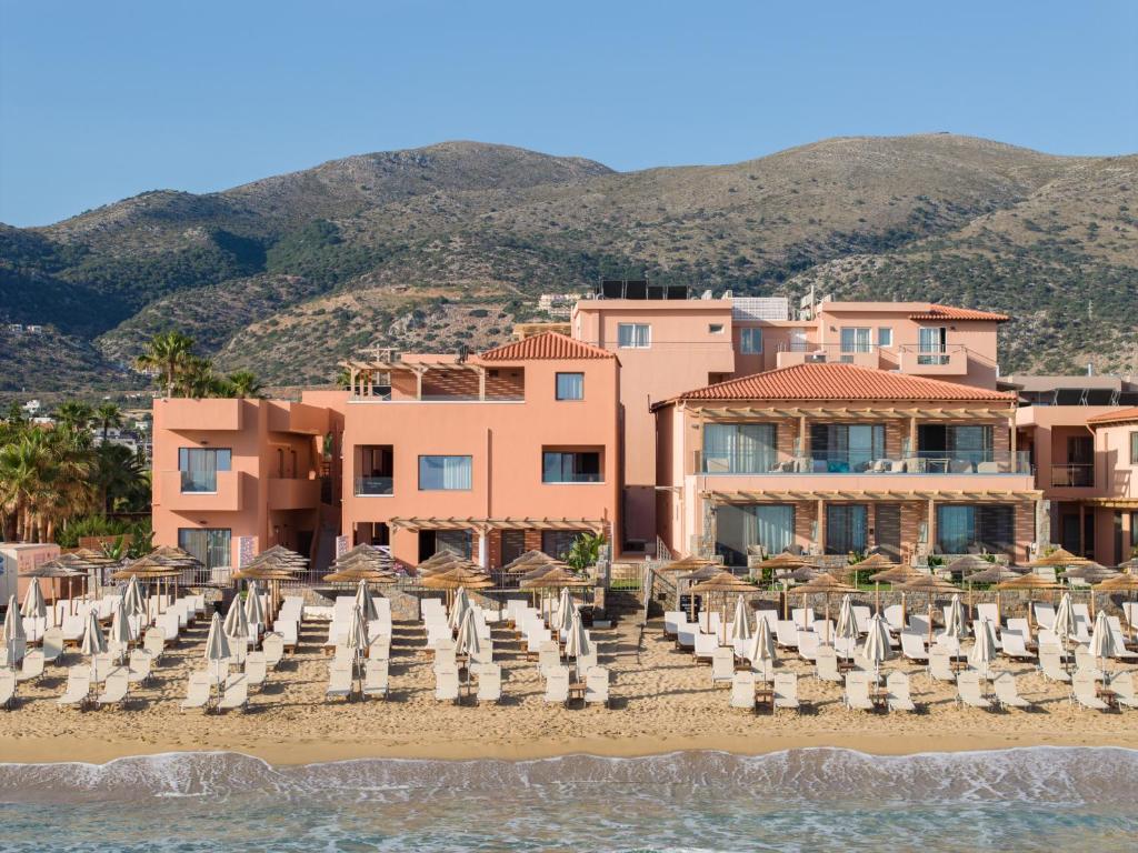 Rozā krāsas viesnīca pie pludmales, ar kalniem fona, kurā redzami saulē iedegti pludmales krēsli un sauļošanās krēsli.