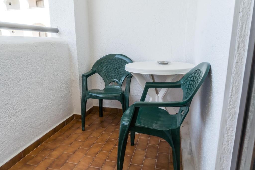 Balkons ar divām zaļām plastmasas krēsliem un baltu galdu, kurā atrodas pelnu trauks. Apkārtējās sienas ir baltas, un grīda ir izklāta ar brūnām flīzēm.
