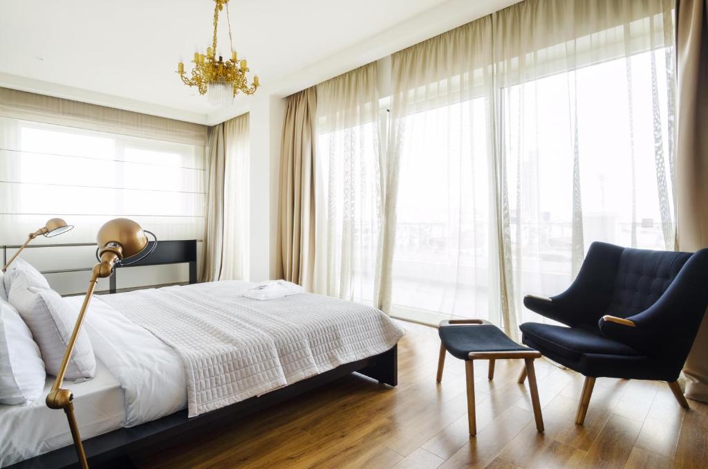 Mūsdienīgs guļamistabas interjers ar gultu, melnu krēslu un elegantu lustru, kas piepilda telpu ar gaismu un komfortu. Lieli logi ar gaisīgām aizkariem nodrošina dabīgu apgaismojumu.
