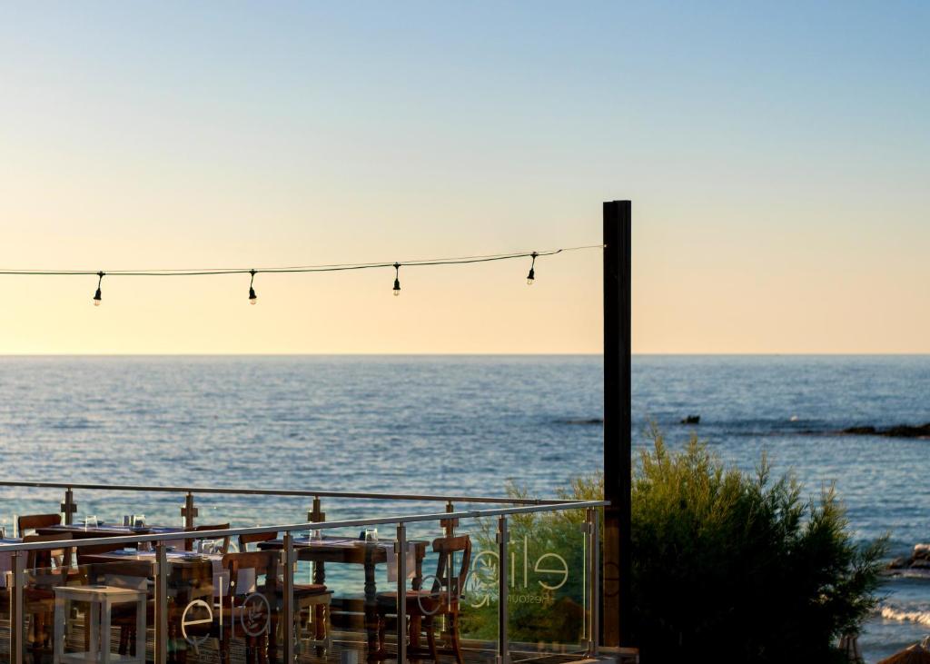 Jūras ainava ar restorāna terasi un gaismas virteni, kas piekarināta virs. Kārtīga galdu izvietojuma un mierīga jūras ūdens virsma.