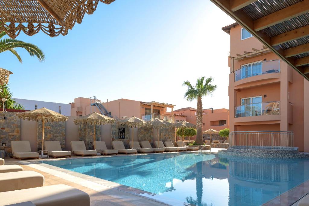 Luksusa viesnīcas baseins ar sauļošanās krēsliem un palmu kokiem, kas rada relaksējošu atmosfēru. Apburošs skats uz ēkām un skaidru debesu fonu.