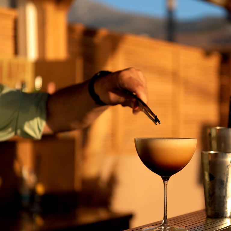 Kokteiļa pagatavošana ar kafijas krēmu, kas tiek dekorēts ar pinceti. Fons ir koka struktūra, kas rada siltu atmosfēru.