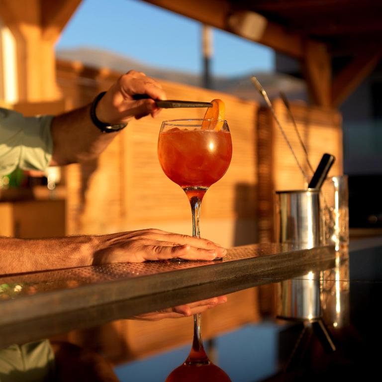 Kokteiļa pagatavošana ar apelsīnu šķēli, kas novietota uz glāzes, atspoguļojot saulrieta gaismu.