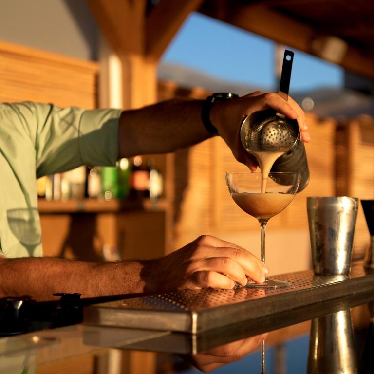 Bārmenis ielej kokteili martini glāzē, izmantojot smalcinātāju, kamēr saule apgaismo bāra vidi.