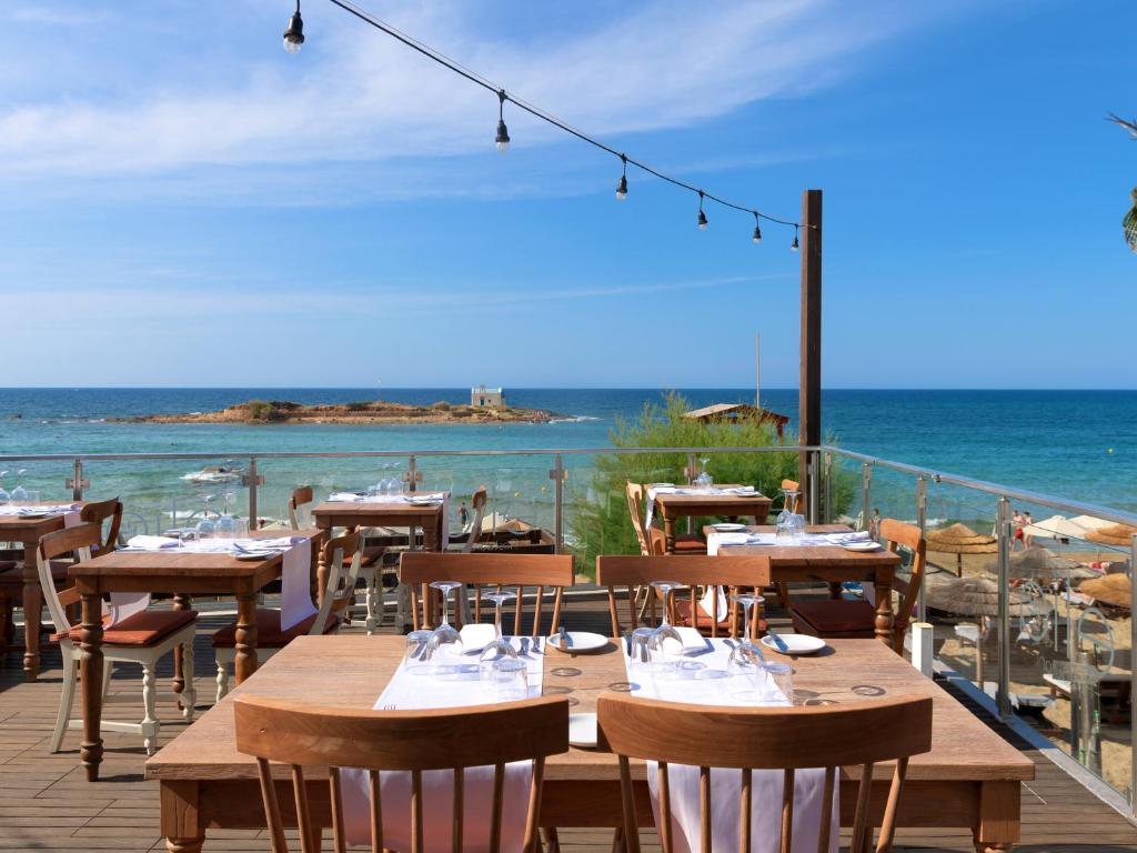 Restorāna terase ar koka galdiem un krēsliem, kas piedāvā skatu uz jūru un pludmali, zem saulainas debesu. Ideāla vieta relaksācijai un baudīšanai pie ūdens.