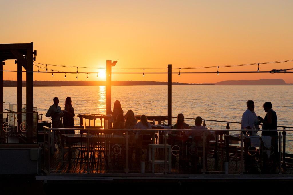 Saulriets pār mierīgu jūru, cilvēki bauda vakaru ārpusē restorānā ar skatu uz ūdeni. Apgaismojums un atmosfēra rada relaksējošu noskaņu.