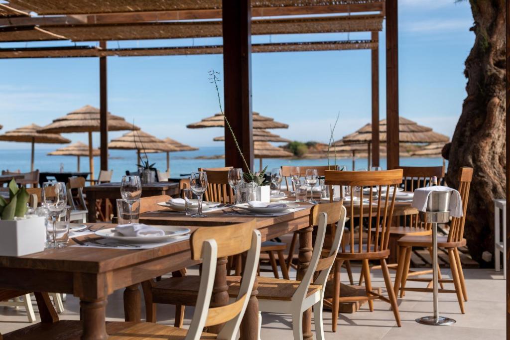 Restorāna terase ar skatu uz jūru, kur izvietoti koka galdi un krēsli, dekorēti ar galda piederumiem un ziediem. Fona ainavā redzami saulessargi un skaidrs debesis.