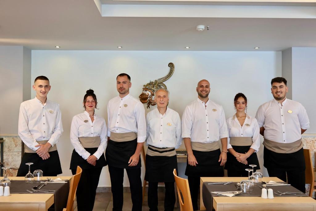Restorāna darbinieki stāv rindā, ģērbušies baltās kreklos un tumšās biksēs, ar smaidiem sejās. Fons ir moderns restorāns ar galdiem un galda piederumiem.