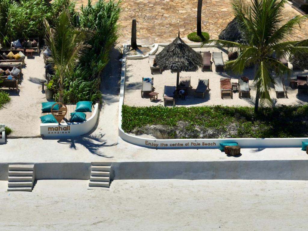 Pludmales atpūtas zona ar šūpuļtīkliem un sauļošanās krēsliem, ko ieskauj tropu augi. Redzama zīme ar tekstu "Enjoy the centre of Paje Beach" un "mahali Zanzibar" logo.