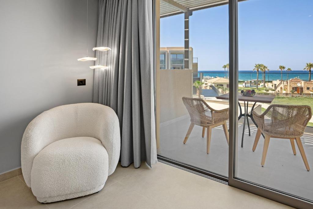 Mūsdienīgs interjers ar baltu krēslu un skatu uz jūru caur stikla durvīm, kur redzami divi vītni krēsli pie galda un palmām apkārt.