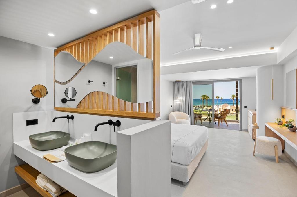 Mūsdienīgs viesnīcas numurs ar elegantu vannas istabu, kurā ir divas zaļas izlietnes, un skatu uz jūru caur plašiem logiem. Interjers ir gaišs un stilīgs, apvienojot koka elementus ar modernām iekārtām.