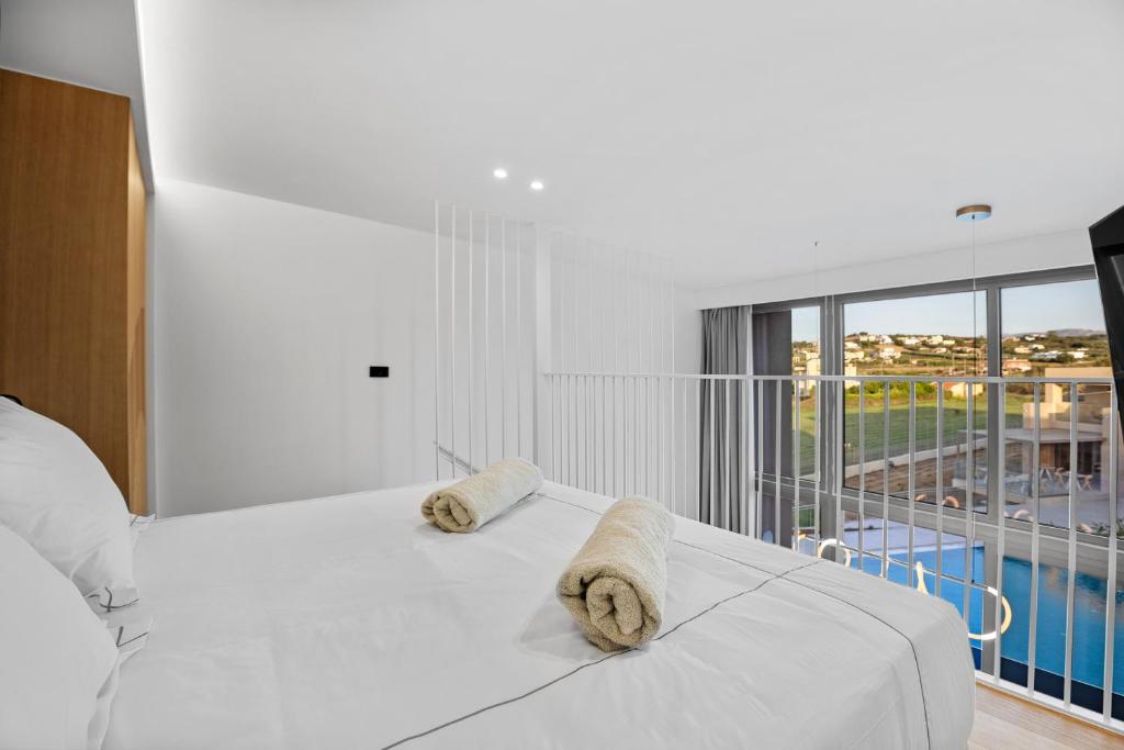 Mūsdienīgs guļamistabas interjers ar baltu gultu un dvieļiem, liela loga ar skatu uz āru un moderna apgaismojuma elementiem. Telpa ir gaiša un plaša, radot komfortablu atmosfēru.