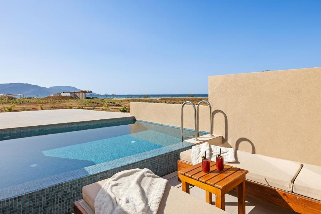 Mūsdienīga atpūtas zona ar privātu baseinu, kurā ir ūdens, un ērta sauļošanās zona ar koka galdiņu un dzērieniem. Fons veido jūras ainava un kalni zem skaidras debess.