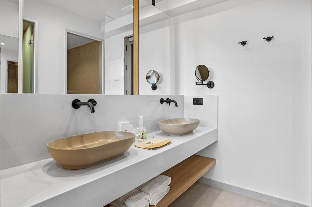 Mūsdienīgs vannas istabas interjers ar diviem sudraba izlietnēm, melniem jaucējiem un spoguļiem. Balta siena un koka plaukts piešķir telpai elegantu izskatu.