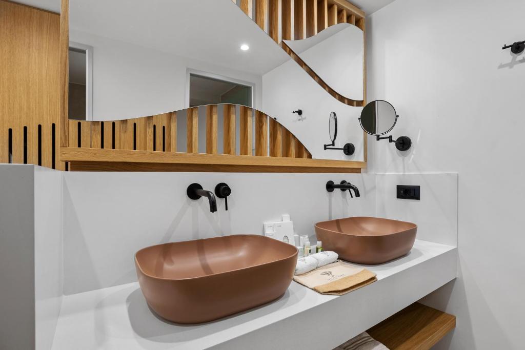 Mūsdienīgs vannas istabas interjers ar divām brūnām izlietnēm, koka elementiem un stilīgu spoguli. Ērta un eleganta telpa ar higiēnas piederumiem uz plaukta.