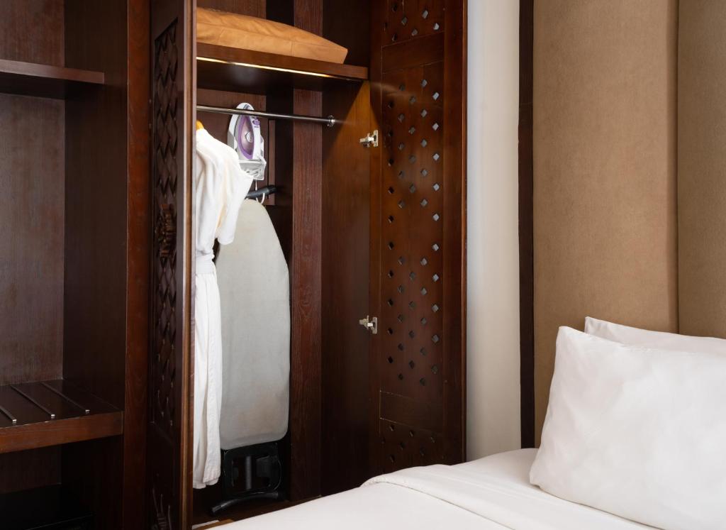 Koka drēbju skapja iekšpuse ar apmetni un dzelzi, blakus gultai ar baltām spilveniem. Ideāla telpa viesnīcas komfortam.