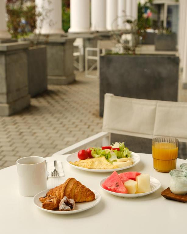 Broka veida galds ar krēmveida kafiju, kruasānu, omleti, dārzeņu salātiem un augļu šķīvjiem, kas novietots uz terases ar kolonnām fonā.