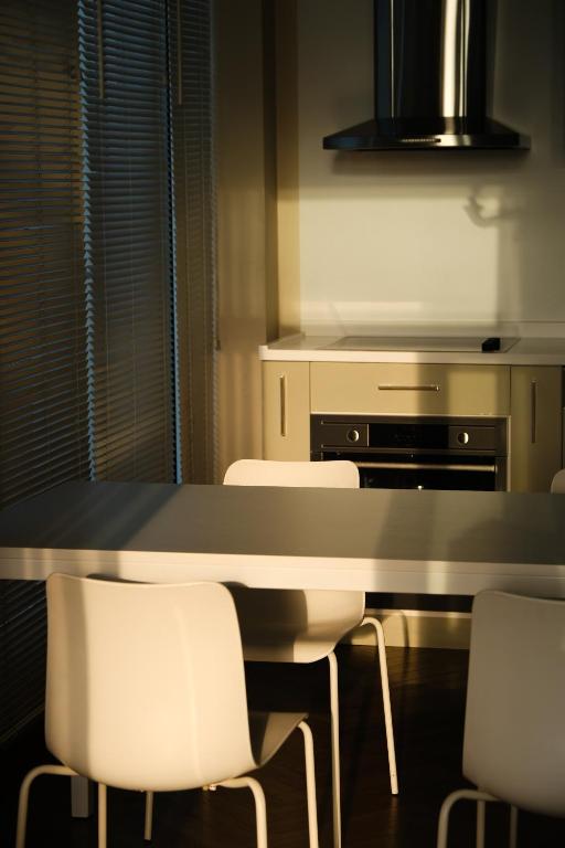 Mūsdienīga virtuve ar baltu galdu un krēsliem, saules gaisma izceļ telpas elementus. Virtuvē ir moderns plīts un izlietne.