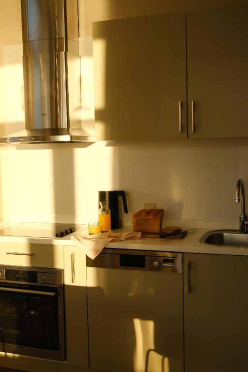 Saules gaismas piepildīta moderna virtuve ar baltām mēbelēm, uz galda ir oranžs dzēriens un maize.