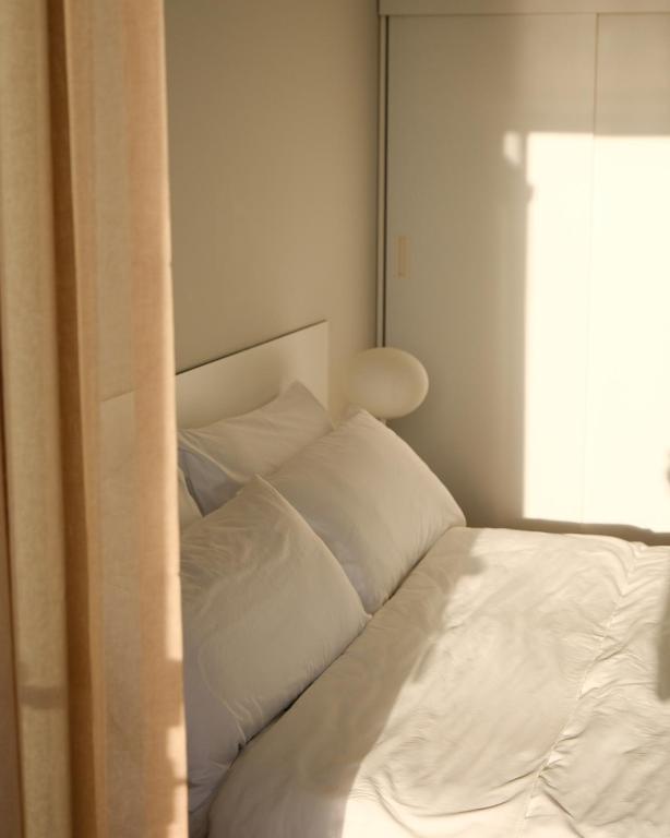 Gaiša guļamistaba ar baltām gultas veļām un mīkstu apgaismojumu, kas ieplūst caur logu. Gultas galvgalis ir vienkāršs, un telpā ir maigs, relaksējošs noskaņojums.