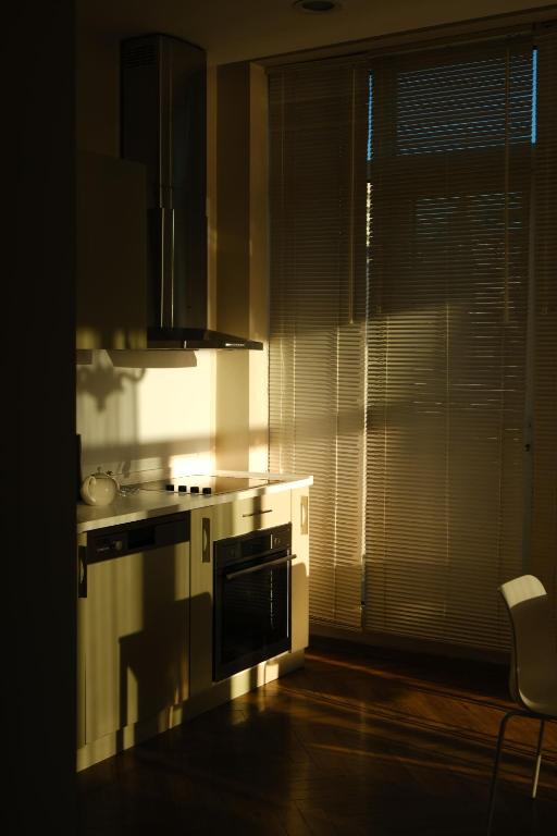 Saulainā virtuve ar gaismu un ēnām, kur redzama modernā iekārtojuma plaukti un galda virsma. Gaismas spēles radītas ar horizontālajām žalūzijām.