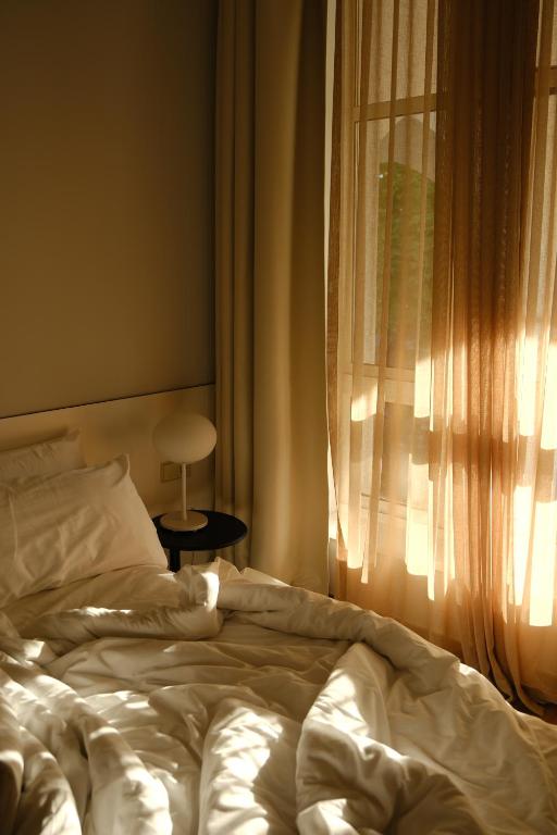 Gaismas piepildīta guļamistaba ar neizklātām gultas veļām un logu ar caurspīdīgām aizkariem. Detaļas rada mierīgu un relaksējošu atmosfēru.