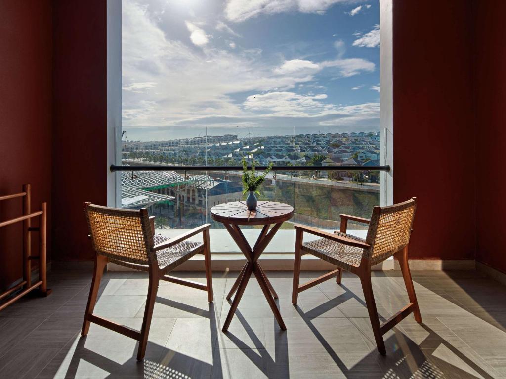 Skats no balkona ar divām koka krēslām un apaļu galdu, kas rotāts ar ziedu podiņu, nodrošina mierīgu un relaksējošu atmosfēru. Aiz loga redzami dzīvojamie rajoni un skaidra debess.