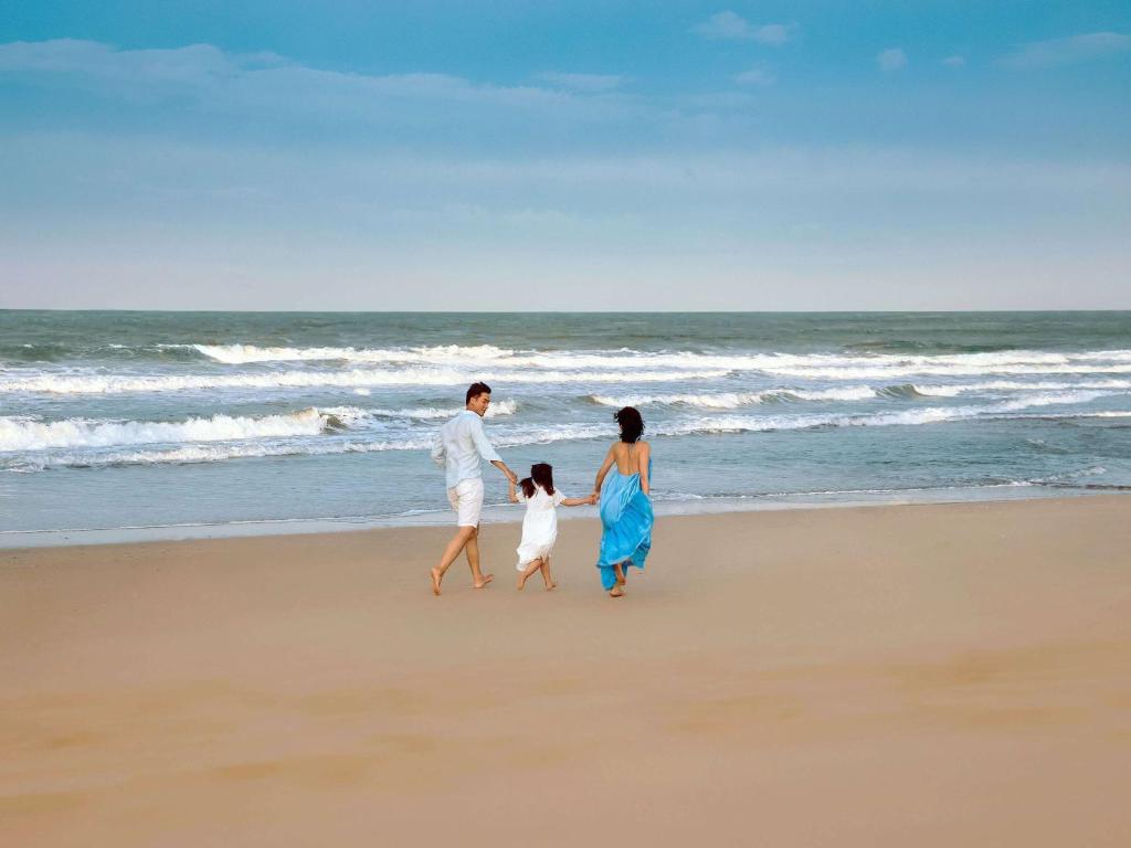 Ģimene pastaigā pa pludmali, baudot saulainu dienu un jūras viļņu skaņas. Bērns tur rokās pieaugušā, kamēr otra pieaugušā kleita plūst vējā.