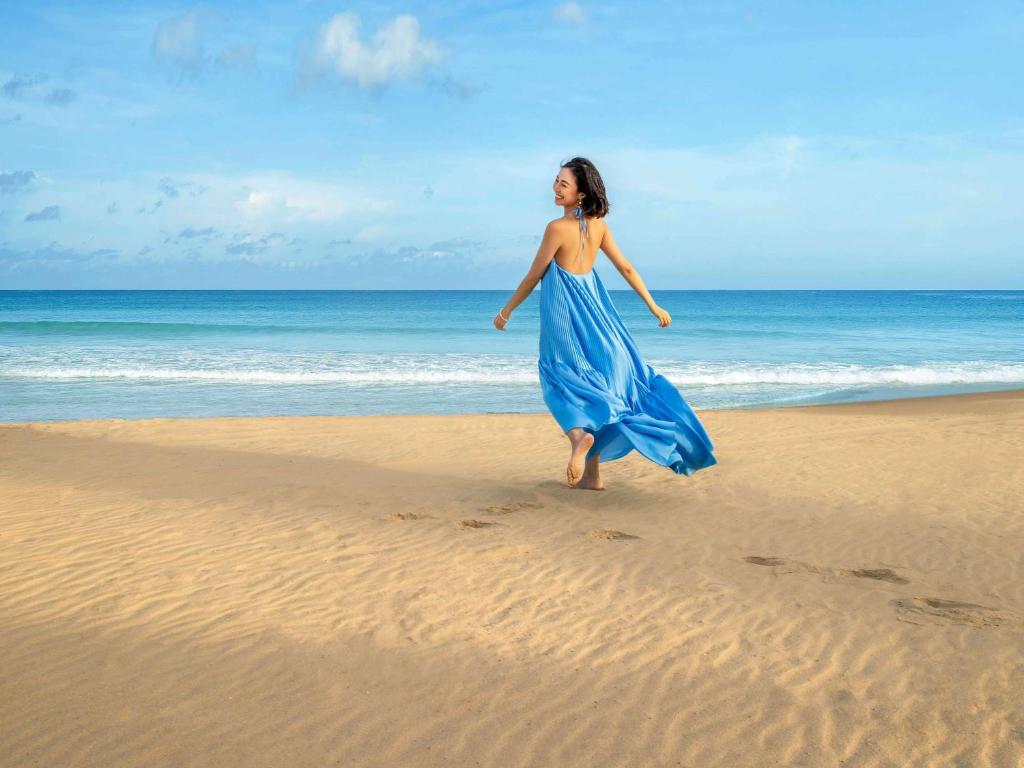 Sieviete zils kleitā soļo pa pludmali, ar jūru un debesīm fonā. Vēja viļņoti mati un kleita piešķir brīvības sajūtu.