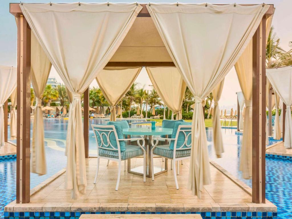 Eleganta atpūtas zona pie baseina ar baltiem aizkariem un zilām mēbelēm, kas piedāvā relaksējošu atmosfēru. Fonā redzama baseina un zaļo palmu ainava.