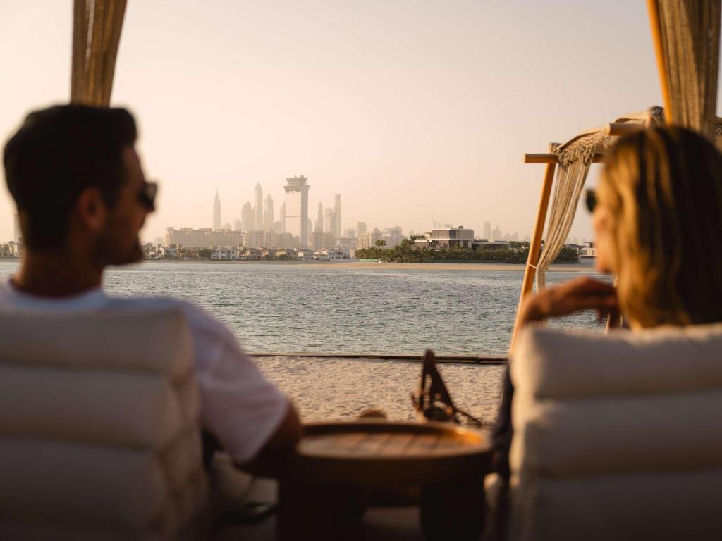 Skats uz Dubaijas panorāmu no pludmales, kur divi cilvēki atpūšas, baudot saulrietu. Klusa atmosfēra ar pludmales ērtībām un ūdens tuvumā.