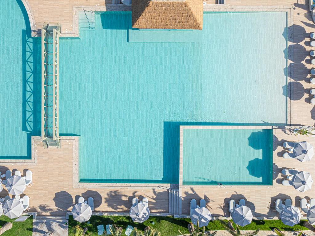 Lielas peldbaseina ar saules gultām un lietus aizsargiem, redzama no augšas. Apkārt baseinam ir koka klājums un zaļie augi.