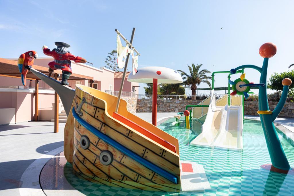 Krāsains bērnu rotaļu laukums ar pirātu tēmu, kurā redzama liela laiva un slidkalniņi. Apkārt ir palmām un saulaina debess, radot priecīgu un jautru atmosfēru.