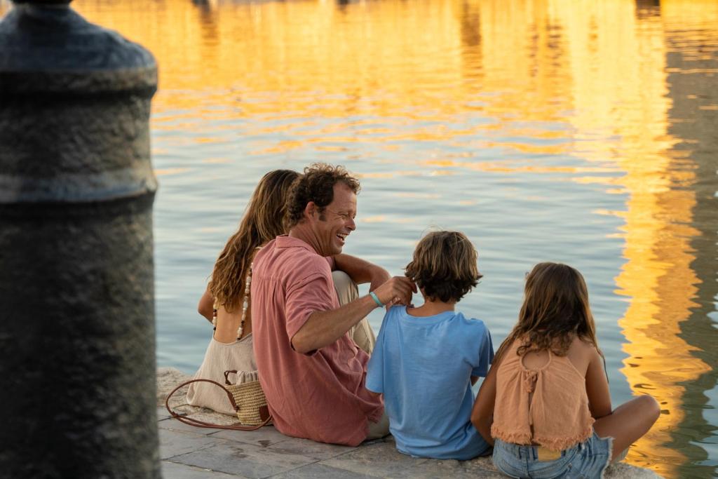 Priecīgas ģimenes brīdis pie ūdens, kur tēvs sarunājas ar bērniem, kam apkārt ir saulrieta gaisma un ūdens atspulgi.