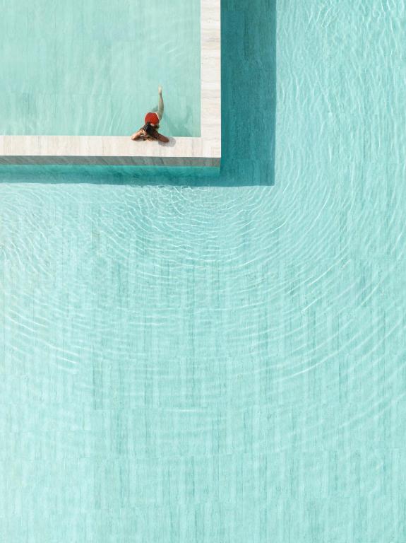 Peldbaseins ar skaidru zilu ūdeni, kurā atrodas cilvēks ar sarkanu peldkostīmu, atpūšoties uz malas. Ūdenī redzami viļņi, kas veido patīkamu un relaksējošu atmosfēru.