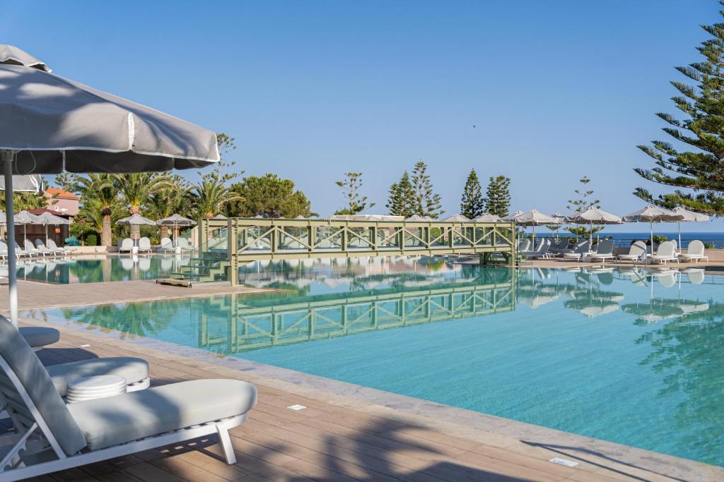 Baseins ar saulessargiem un palmām, pie kura ir tiltiņš, piedāvā relaksējošu atmosfēru tropiskā vidē. Skats uz ūdens virsmu un zilo debesu fonu rada mierīgu noskaņu.