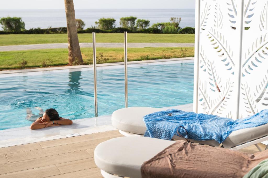Atpūta pie baseina ar skatu uz jūru, kur cilvēks relaksējas, balstoties uz baseina malas. Apkārt ir zaļi zālieni un brīnišķīga atmosfēra.