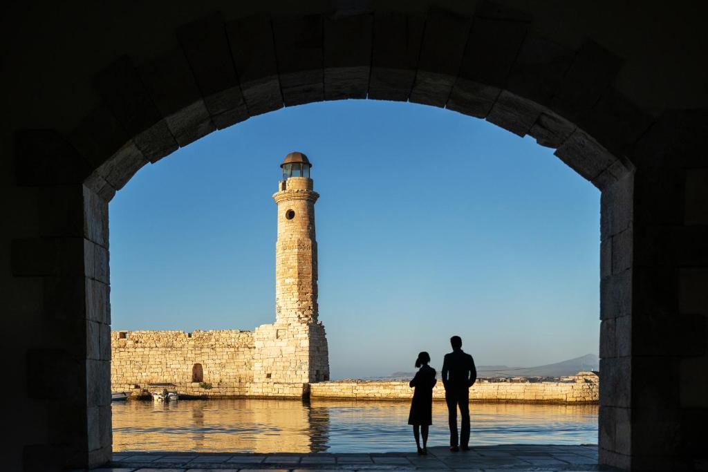 Skats caur arku uz gaismas bāku pie ūdens, kur stāv divi cilvēki. Fons ir skaidrs debesis un akmens mūris, kas rada romantisku atmosfēru.