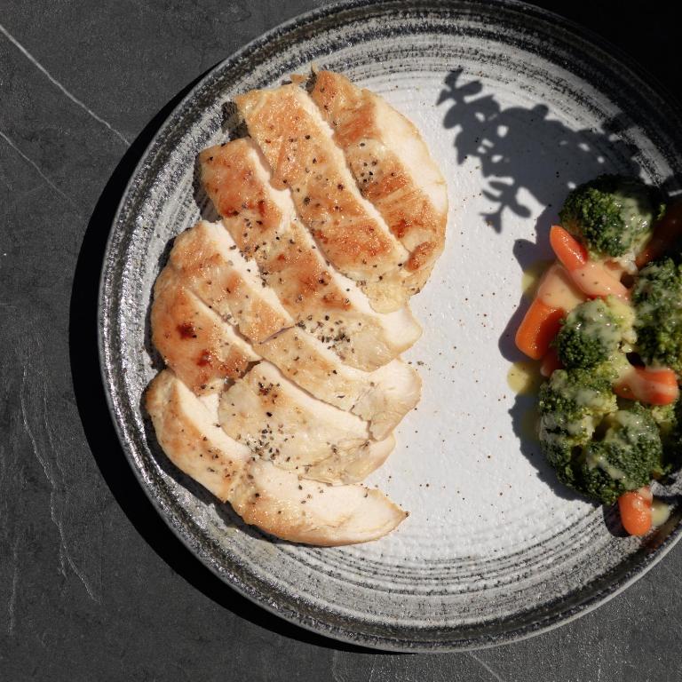 Grilēta vistas fileja ar garšvielām, pasniegta kopā ar dārzeņu maisījumu, kas satur brokoļus un burkānus. Plāksne ar modernu dizainu uz tumša fona.