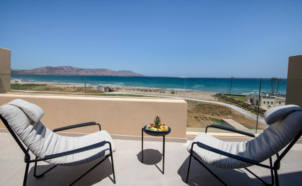 Balkona skats uz pludmali ar divām ērtām krēsliem un mazu galdiņu, uz kura ir augļu šķīvis. Fonā redzams zils jūras ūdens un kalnu ainava.