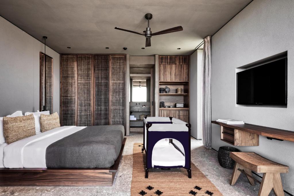 Mūsdienīgs guļamistabas interjers ar koka mēbelēm, pelēku gultu un bērnu gultiņu, kas radīts relaksējošai atmosfērai. Sienas ir gaišas, un telpā ir televizors un eleganta apgaismojuma sistēma.